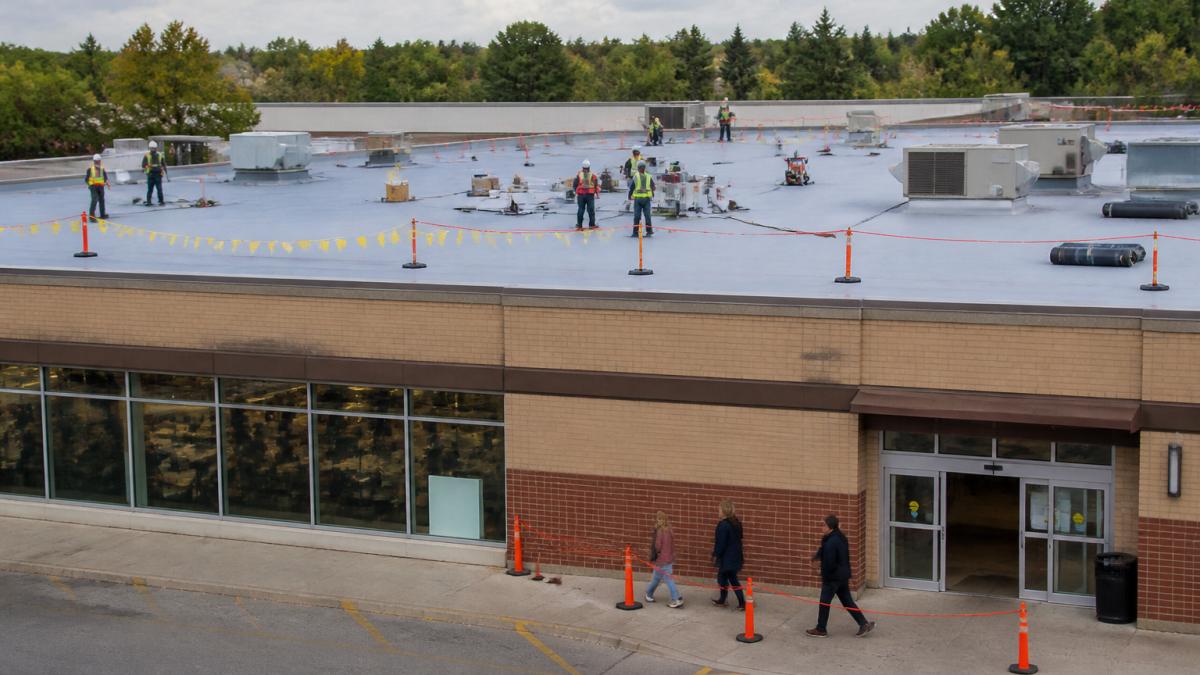 Roofing crew performing phased roof repairs while customers enter a retail store below marked by safety barriers.